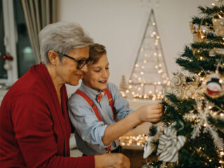 A woman helps a young boy put an ornament on a Christmas tree.