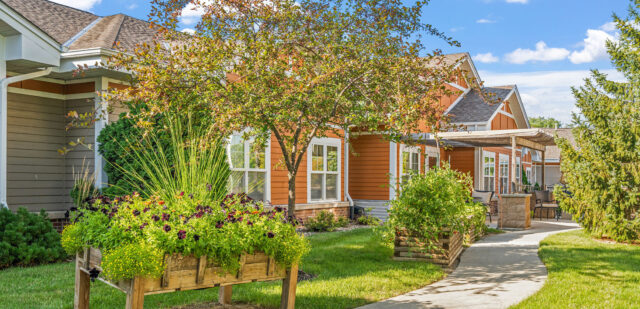 This is a picture of Newton Village's garden, featuring raised planters with vibrant flowers, a winding concrete path and a seating area under a pergola. The building has warm-toned siding and large windows.