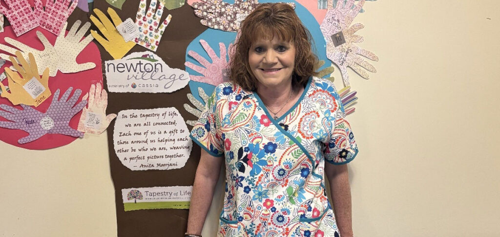 Janette, a CNA at Newton Village, smiles in front of a colorful handprint mural celebrating connection and care in the senior living community.