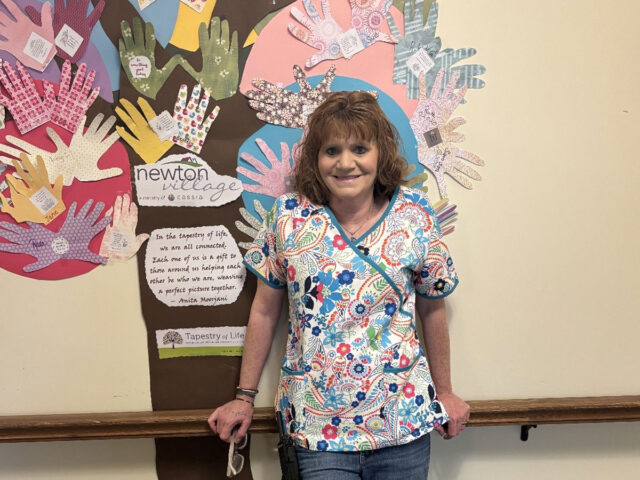 Janette, a CNA at Newton Village, smiles in front of a colorful handprint mural celebrating connection and care in the senior living community.
