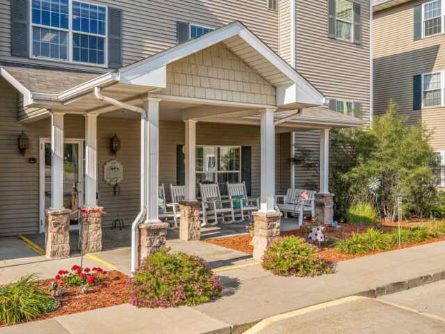 Front entrance of Newton Village Assisted Living with rocking chairs, landscaped flowers, and welcoming porch.