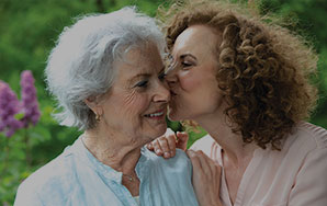Two older adults sharing a close moment outdoors with one leaning in toward the other, representing compassionate memory care and family support like at Newton Village