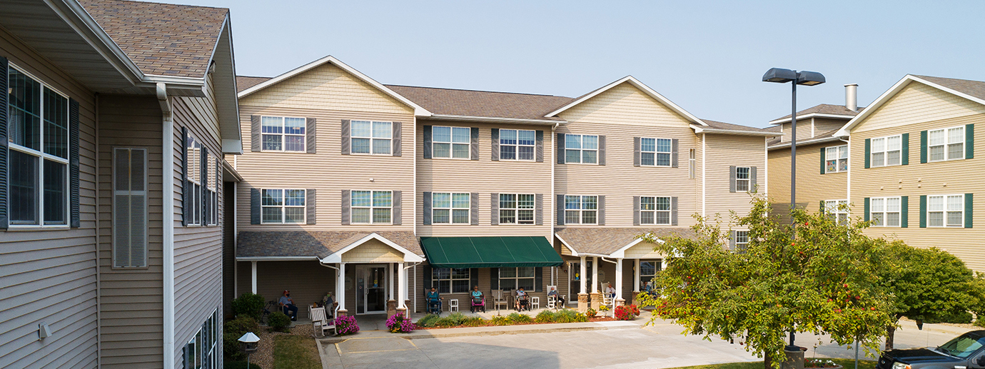 Exterior view of Newton Village showing the main building entrance with covered entryways, landscaping, and residents seated along the front