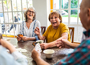 Group of older adults playing cards together at a table in a bright living room with large windows, representing social activities and community life like at Newton Village