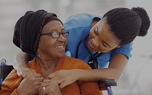 Caregiver assisting an older adult seated in a wheelchair during a supportive interaction, representing long-term care services like at Newton Village