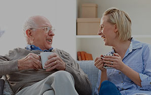 Two older adults seated together on a couch holding coffee mugs and talking in a home-like setting, representing assisted living support and companionship like at Newton Village