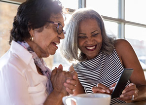 Two older adults seated at a table with a coffee cup, looking at a phone together in a bright setting, representing social interaction and connection like at Newton Village