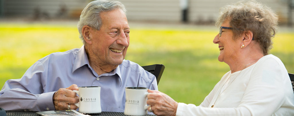 Two older adults seated outdoors holding coffee mugs and talking at a table, representing social interaction and community living like at Newton Village