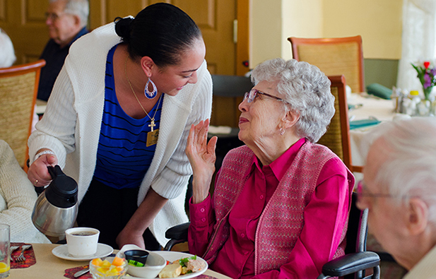 Staff member serving coffee to an older adult seated in a wheelchair during a meal in a dining setting, representing supportive dining care like at Newton Village