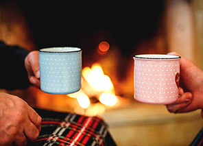 Two adults holding mugs near a fireplace with a warm fire in the background, representing cozy social moments and gathering spaces like at Newton Village