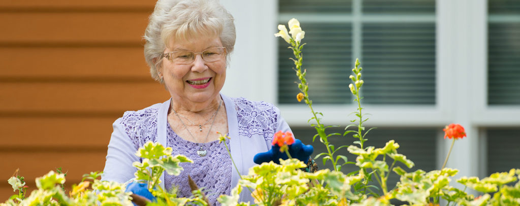 Older adult tending to flowers in a garden bed outside a residential building, representing gardening and outdoor activities like at Newton Village