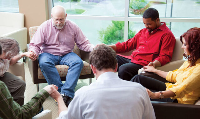 Group of adults seated in a circle holding hands in a shared space near large windows, representing spiritual care and group support like at Newton Village