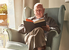 Older adult seated in a cushioned armchair reading a book near a window with natural light, representing quiet activities and relaxation like at Newton Village