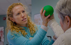 Staff member assisting an older adult with a hand exercise using a therapy ball in a clinical setting, representing short-term rehabilitation services like at Newton Village