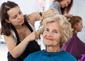 Staff member styling an older adult’s hair in a salon chair with mirrors and other clients in the background, representing personal care services like at Newton Village